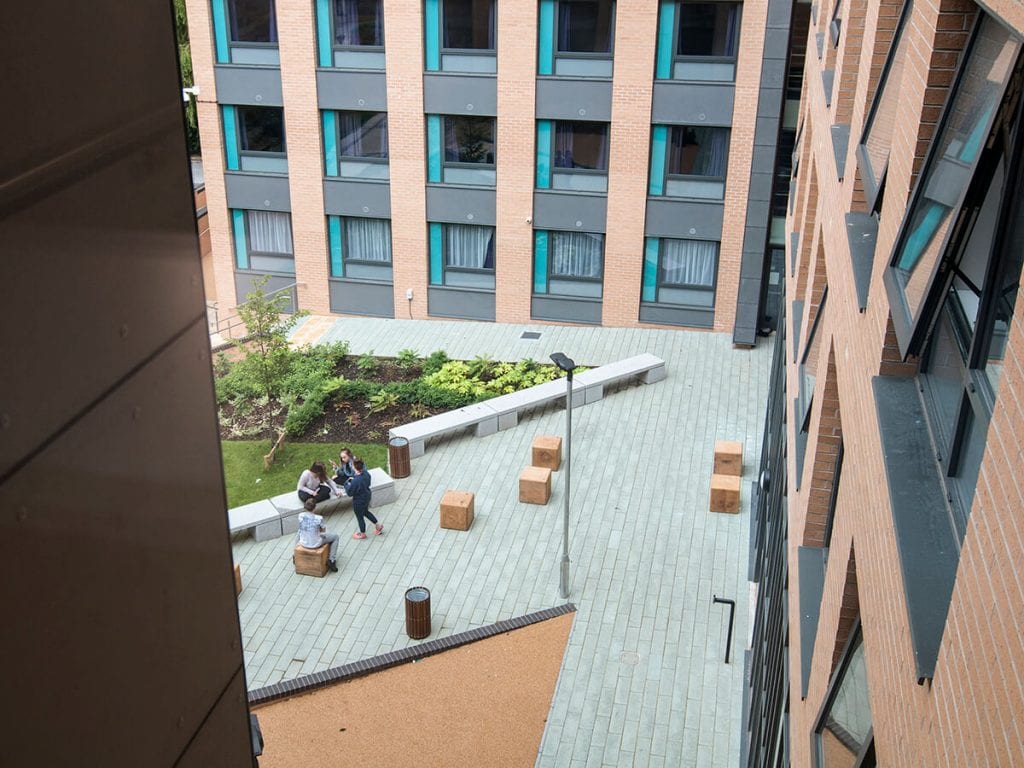 Natural Dimensions View of Fountians court Leeds Trinity University with linear granite bench, individual timber bale seats, plank paving carpinus betulus trees, fatsia japonica and buff resin bound gravel on ramp