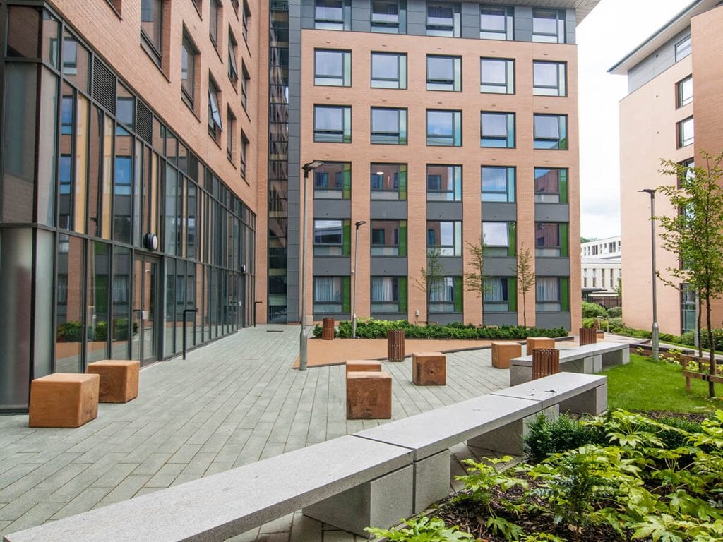 View of Fountians court Leeds Trinity University with linear granite bench, individual timber bale seats, plank paving carpinus betulus trees, fatsia and buff resin bound gravel on ramp